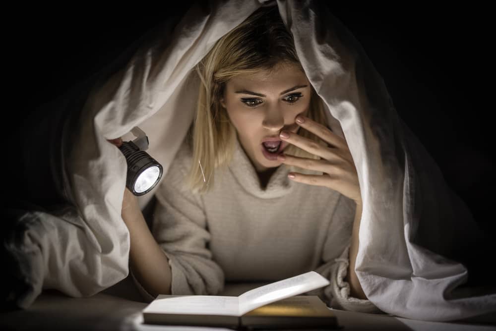 Joven aterrorizada leyendo uno de los mejores libros de terror en la cama, iluminando las páginas con una linterna.