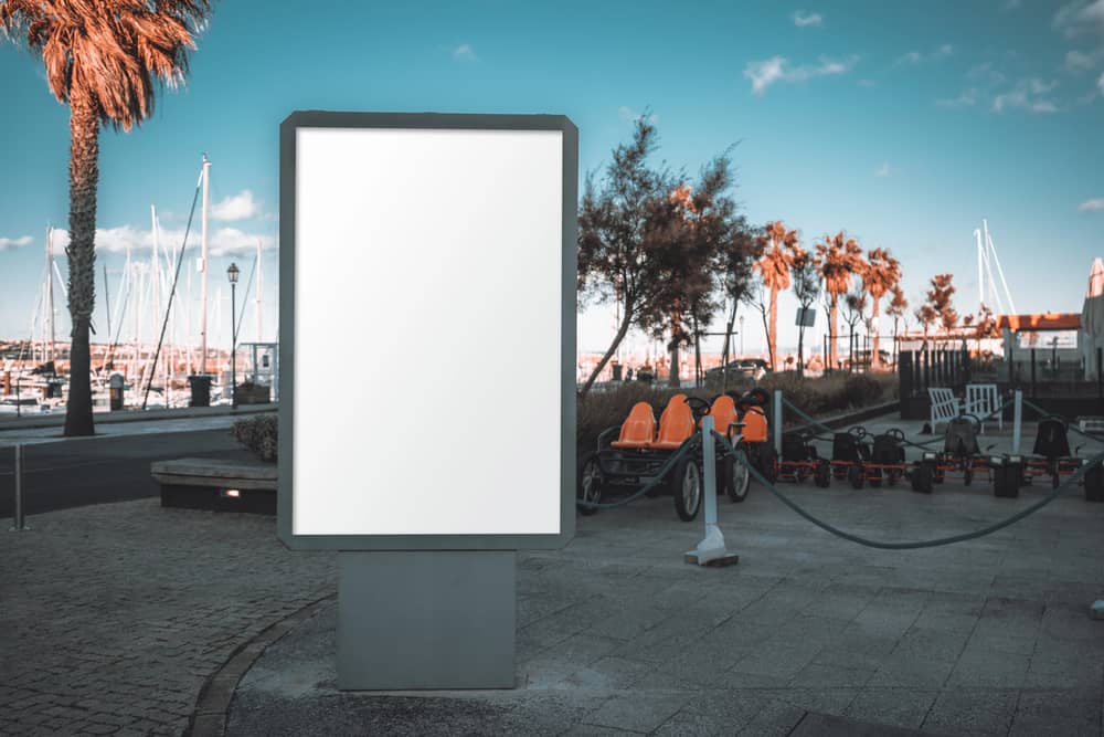 Fuentes de Ingresos Adicionales: ¿En Qué Puedes Ganar Dinero Extra? 7 Una valla publicitaria vertical en blanco, ubicada en una zona portuaria con palmeras y botes al fondo, ideal para publicidad o como fuente de ingresos pasivos.