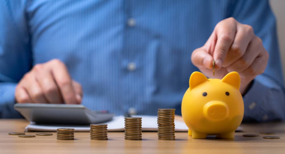 Persona en camisa azul calculando con una calculadora y depositando una moneda en una alcancía de cerdito amarilla, con pilas de monedas en el escritorio, simbolizando el ahorro y la elaboración de un presupuesto.