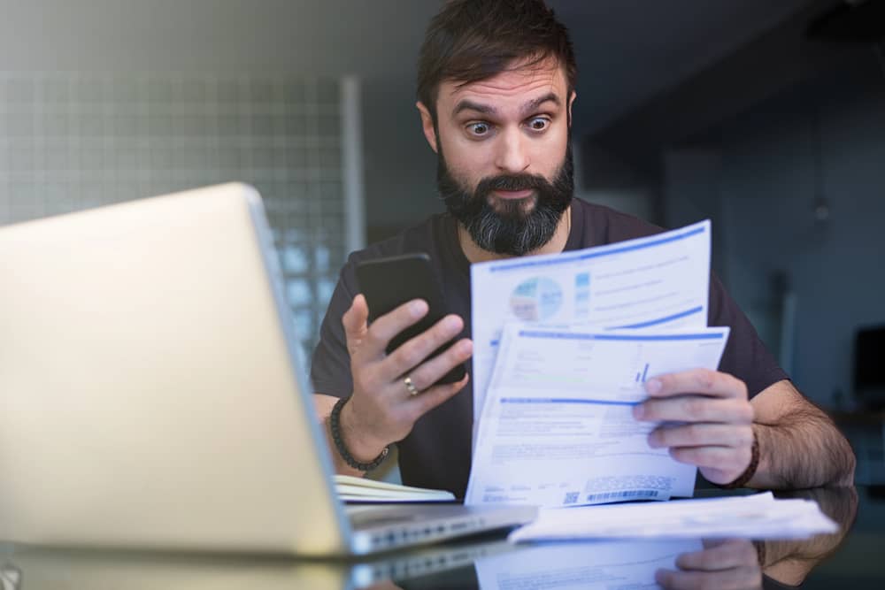 Un hombre con barba y expresión de sorpresa mira documentos financieros y un teléfono móvil, con una laptop al fondo, representando la complejidad de gestionar un presupuesto como freelance.