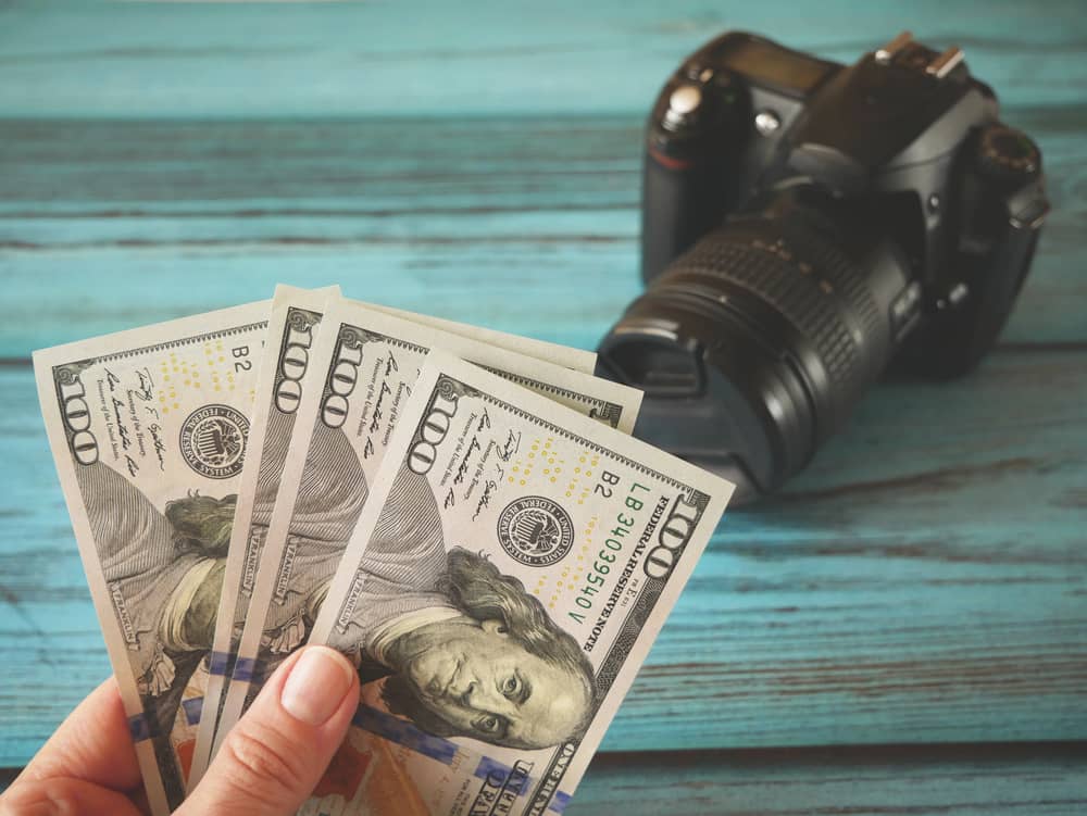 Fuentes de Ingresos Adicionales: ¿En Qué Puedes Ganar Dinero Extra? 6 Una mano sostiene varios billetes de cien dólares, con una cámara DSLR profesional en el fondo sobre una superficie de madera azul, simbolizando cómo la fotografía de stock puede ser una de las mejores fuentes de ingresos adicionales.