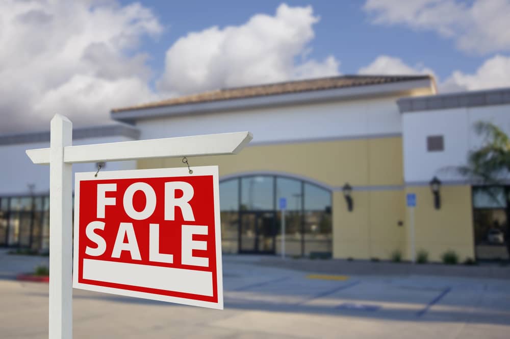 Fuentes de Ingresos Adicionales: ¿En Qué Puedes Ganar Dinero Extra? 9 Un cartel de "For Sale" (En Venta) frente a un edificio comercial moderno, bajo un cielo parcialmente nublado.