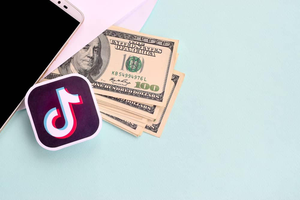 Fuentes de Ingresos Adicionales: ¿En Qué Puedes Ganar Dinero Extra? 5 Un smartphone, billetes de cien dólares y un logo de TikTok sobre un fondo azul claro, representando el potencial de ganar dinero a través de las redes sociales.