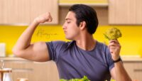 Hombre sonriente flexionando el bíceps mientras sostiene un brócoli en una cocina, representando fuerza y salud.